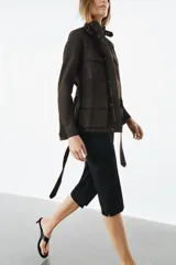 Chaqueta de estilo safari confeccionada en mezcla de algodón con acabado encerado. Presenta cuello alto con trabilla ajustable y hebilla metálica, cierre frontal con cremallera y botones ocultos bajo solapa, bolsillos de plastrón con solapa en el delantero y cinturón ajustable del mismo tejido para anudar en la cintura.