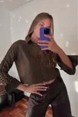 Camisa corta marrón de cuero ovino desgastado, con cuello clásico, cierre frontal con botones, mangas largas con cutouts y puños anchos dorados.