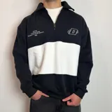 Buzo de algodón con medio cierre frontal y cuello tipo polo. Presenta un diseño de bloques de color en tonos gris oscuro y blanco, con bordados decorativos en el pecho.