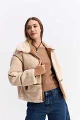 Campera tipo teddy de textura suave y acabado de peluche en color beige. Presenta cuello amplio con detalle de hebilla, cierre frontal y recortes en contraste de color marrón que marcan la estructura de la prenda.