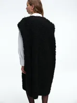 Chaleco largo reversible de diseño versátil. Por un lado, presenta una textura de peluche sintético en color marrón, y por el otro, un acabado liso. Cuenta con cierre frontal de botones y corte recto.