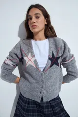 Cardigan corto tejido en color gris, con escote en V y cierre frontal de botones. Presenta un diseño estampado con estrellas y líneas geométricas en tonos oscuros y rojizos, con mangas largas y puños acanalados.