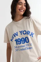 Remera de manga corta color beige, con estampa frontal en tono azul con la leyenda "New York 1990", de corte holgado y diseño casual.