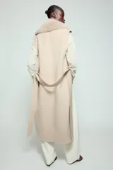 Chaleco largo de mezcla de lana color beige, de corte recto con manga sisa. Cuenta con cuello solapa con aplicación de pelo sintético extraíble, bolsillos laterales ocultos y un cinturón ajustable del mismo tejido para anudar en la cintura.
