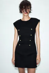 Vestido corto de estilo militar en color negro, con cuello redondo, hombreras y manga sisa. Presenta un diseño frontal con falsos botones dorados dispuestos en filas horizontales. Cierre lateral con cremallera oculta y abertura en la espalda con botón.