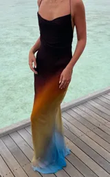 Vestido largo de seda o material similar, con tirantes finos y escote drapeado tipo cowl neck. Presenta un efecto degradado de color que va del negro/marrón oscuro en el busto, pasando por tonos naranjas y amarillos, hasta terminar en un azul vibrante en el ruedo.