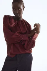 Blusa de satén color borgoña con mangas globo largas y puños anchos. Presenta un detalle de drapeado o nudo en el centro del escote redondo.