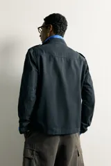 Chaqueta estilo militar relaxed fit de color azul marino, confeccionada en mezcla de algodón y lino. Presenta cuello subido, manga larga con puño abotonado, hombros con trabillas y cuatro bolsillos delanteros de parche con solapa. Se cierra al frente con botonadura metálica dorada.
