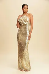 Vestido largo de fiesta totalmente cubierto de lentejuelas doradas, con escote halter y un detalle de flor grande tridimensional en el pecho. Presenta una silueta ajustada que se extiende hasta el suelo.