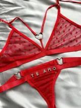 Conjunto de lencería rojo de tul con diseño de corazones aterciopelados en relieve. Incluye un soutien tipo triángulo con argollas metálicas en forma de corazón y tiras regulables, y una bombacha colaless con tiras laterales ajustables unidas por argollas en forma de corazón y el texto "TE AMO" aplicado con apliques de pedrería brillante.