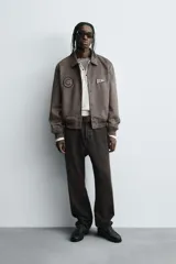 Cazadora estilo varsity de tejido efecto ante en color beige. Presenta cuello solapa, cierre frontal con botones a presión y puños y ruedo con acabado en rib a rayas negras. Incluye parches bordados decorativos en el pecho y la manga, además de bolsillos laterales de doble vivo.