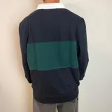 Buzo tipo polo de rugby con cuello blanco, manga larga y puños acanalados. Presenta un diseño de bloques de color en azul marino y verde, con un pequeño logo bordado en el pecho.