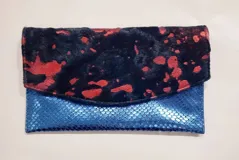 Sobre de mano Carolina De Cunto, rectangular, con solapa y cierre de imán. Combinación de cuero con textura simil piel de serpiente en color azul metálico en la base y cuero con pelo estampado con manchas en tonos negros y rojos en la solapa.
