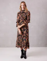 Vestido midi de gasa con estampado de arabescos en tonos cálidos sobre fondo negro. Presenta cuello alto con detalle de punto smock, mangas largas con puños elásticos y volados, y falda con tajo lateral y ruedo con volado. Incluye forro interno y cierre posterior.