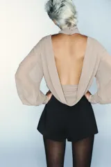 Body de cuello subido con mangas largas abullonadas y puños elásticos. Presenta un diseño combinado con paneles de tela semitransparente en la parte superior y cierre inferior con botones a presión.
