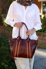 Cartera tipo tote de cuero vacuno en combinación de colores bordó y camel. Presenta un diseño con franjas verticales texturizadas al frente, doble asa de mano y un detalle de borla colgante con argolla metálica en uno de los laterales.