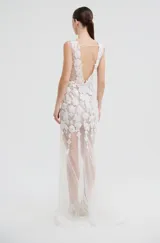 Vestido largo de fiesta con escote en V, confeccionado en tul transparente con aplicaciones de encaje floral blanco. Presenta un diseño sin mangas con detalle de volado en uno de los hombros y falda con caída fluida.