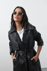 Trench coat de símil cuero color marrón oscuro, con solapas amplias, cierre frontal con botones, cinturón ajustable en la cintura y mangas con tiras ajustables. Presenta un largo por debajo de la rodilla y bolsillos laterales.