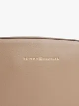 Cartera tipo camera bag de Tommy Hilfiger en color beige, con acabado granulado y logo de la marca en letras doradas en el frente. Cuenta con cierre de cremallera superior, compartimento principal, bolsillo interior abierto y bolsillo interior con cremallera. Incluye una correa ajustable de tejido textil con detalles distintivos de la marca.
