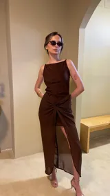 Vestido largo marrón oscuro, ajustado al cuerpo, con escote cuadrado y tirantes finos. La falda presenta un drapeado frontal anudado que crea una abertura alta.