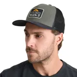 Gorra tipo trucker de cinco paneles con frente y visera en color verde oliva, y parte trasera de malla negra. Presenta un parche frontal rectangular con diseño gráfico de un atardecer y el logo de la marca Roark. Cuenta con cierre ajustable en la parte posterior.