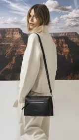 Bandolera de tamaño mediano en color gris, confeccionada en símil cuero con textura granulada. Presenta un bolsillo frontal con cierre, herrajes metálicos oscuros y correa ajustable y desmontable.