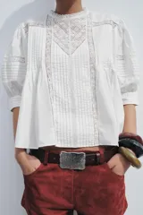 Blusa de algodón color blanco con cuello redondo y mangas abullonadas. Presenta detalles de puntilla a tono en el pecho y las mangas, bajo recto y cierre posterior con abertura y botón.