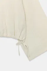 Camisa tipo capa de color beige, con cuello redondo y escote en pico en la espalda. Presenta mangas largas amplias y bajo ajustable con lazada lateral.