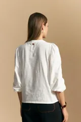 Camisa de algodón blanca con delicadas líneas bordadas verticales y textura calada. Presenta escote redondo con cierre frontal de botones, mangas largas con leve volumen en los hombros y puños elásticos.