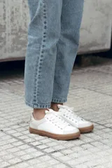 Zapatilla de estilo urbano en tonos marfil, con detalles en color dorado en el talón y ojales metálicos. Presenta suela de goma color caramelo y cordones al tono.