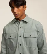 Camisa manga larga de sarga en color verde militar, con cuello inglés, cierre frontal de botones negros y dos bolsillos con solapa en el pecho.