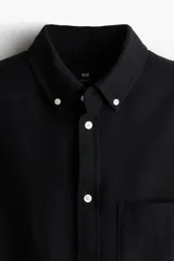 Camisa de algodón estilo Oxford, color negro, con cuello americano abotonado, bolsillo en el pecho y cierre frontal con botones blancos.