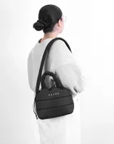 Bolso tipo tote pequeño color negro, con textura acolchada y logo de la marca en el frente. Tiene doble asa corta y correa larga para colgar del hombro.