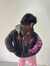 Campera reversible con cierre frontal. Un lado es de algodón negro con estampa y bordado en color rosa, y el otro lado es de tela polar de pelo largo en color gris. Presenta corte oversize, capucha y puños acanalados.