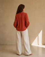 Camisa de lino color terracota, de corte holgado, con cuello camisero, cierre frontal de botones y mangas largas con detalles perforados y trabilla para arremangar.