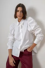 Camisa de algodón color blanco, con cuello clásico, cierre frontal de botones ocultos y mangas largas abullonadas con puños estructurados. Presenta detalles de botones con strass en puños y cuello.