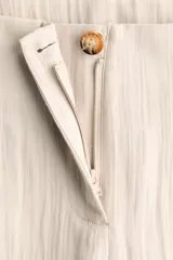 Pantalón tobillero de tiro alto en tejido arrugado color beige, con cierre de botón, bolsillos laterales y bajo ajustable con cordón.