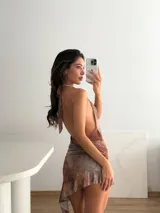 Vestido corto con estampado abstracto en tonos tierra y violetas, escote halter profundo y fruncido en la cintura con un dije de estrella de mar dorado. La falda tiene un ruedo asimétrico con volado.