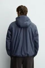 Parka de tejido técnico con acabado engomado, cuello subido y capucha ajustable. Presenta cierre frontal con cremallera oculta bajo solapa con botones a presión, bolsillos laterales de vivo y mangas largas con puños ajustables.
