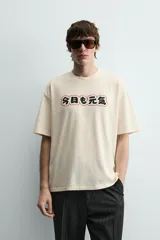 Camiseta relaxed fit color crema, con cuello redondo y manga corta. Presenta un estampado frontal con caracteres japoneses en negro y contorno rojo.