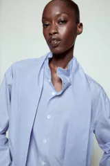 Camisa de algodón color celeste, con cuello alto y lazo integrado en el mismo tejido. Presenta cierre frontal con botones, manga larga con puños y un bolsillo frontal en el pecho. Diseño de corte recto.