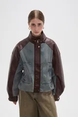 Campera estilo bomber con diseño color block, combinando paneles de denim lavado con mangas y detalles en cuero sintético color marrón oscuro. Presenta cuello alto con tira ajustable, cierre frontal con botones a presión y bolsillos frontales tipo parche.