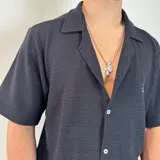 Camisa de manga corta tejida en color negro, con cuello tipo camp y cierre frontal de botones blancos. Presenta un calce holgado y un pequeño logo bordado en el pecho.