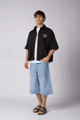 Camisa de manga corta negra, de corte boxy, con cuello camisero, cierre frontal de botones y bordado circular en el pecho.