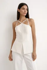Blusa blanca de tweed con diseño estructurado, escote halter y tirantes finos cruzados decorados con pedrería. Presenta un corte entallado en el torso con abertura frontal y silueta peplum en la parte inferior.
