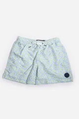 Short de baño celeste con estampado de hojas pequeñas en tonos verdes y blancos. Cuenta con cintura elástica con cordón ajustable, bolsillos laterales y un parche circular con logo en la parte inferior.