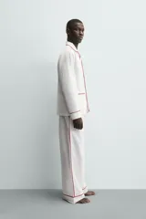 Pijama de dos piezas confeccionado en popelín de algodón blanco, con vivos de ribete en color bordó. Incluye una camisa de manga larga con cuello bowling, bolsillos de plastrón y cierre frontal de botones, y un pantalón de corte relajado con cintura elástica y bolsillos laterales.