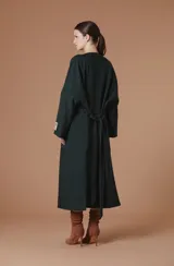 Saco Natalia Antolin de mujer color verde oscuro, de corte largo y diseño relajado con cuello redondo, mangas anchas y cierre frontal de botones oscuros. Incluye una etiqueta decorativa distintiva de la marca en el puño de la manga izquierda.