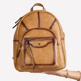 Mochila cartera Korium Vera de cuero sintético en color tostado con efecto envejecido y costuras decorativas. Cuenta con un bolsillo frontal con cierre, detalles de tachas y el logo metálico de la marca.