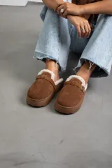 Zueco de gamuza color marrón, con interior forrado en corderito sintético, suela de plataforma plana y detalle de tira con hebilla metálica en el empeine.